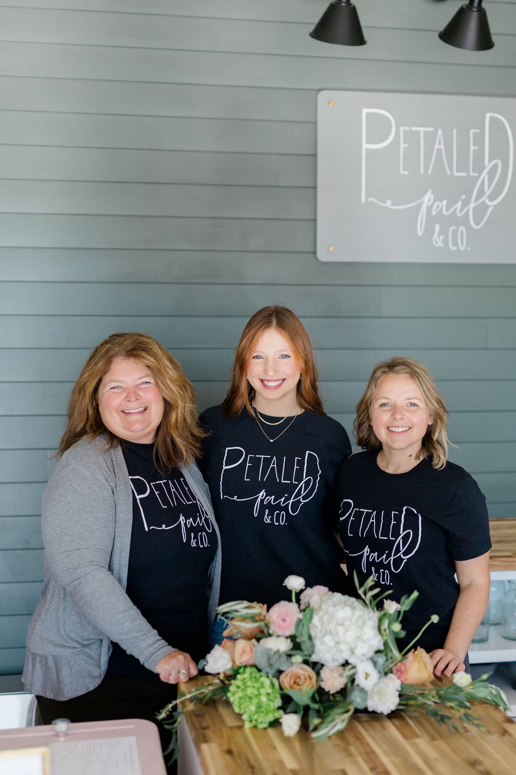 Three smiling women standing behind a table with a flower arrangement, in front of a wall with a sign reading 'Petaled' and 'co.'