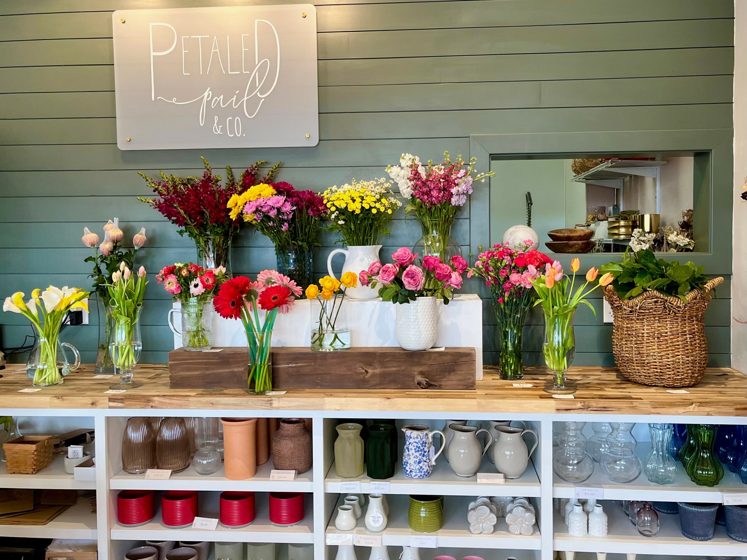Display of colorful flowers in vases on a wooden counter with shelves of cups and bowls below, and a sign in the background.