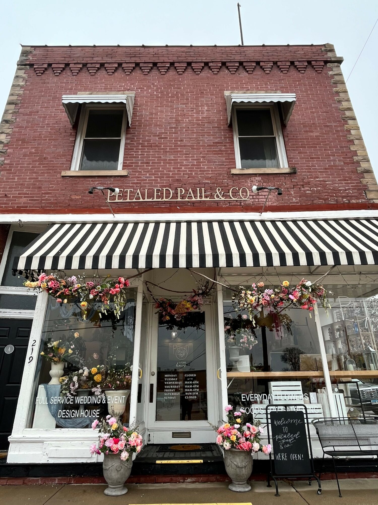 Two-story brick building with striped awning, flower baskets, and glass door entrance, storefront of a flower shop or cafe.