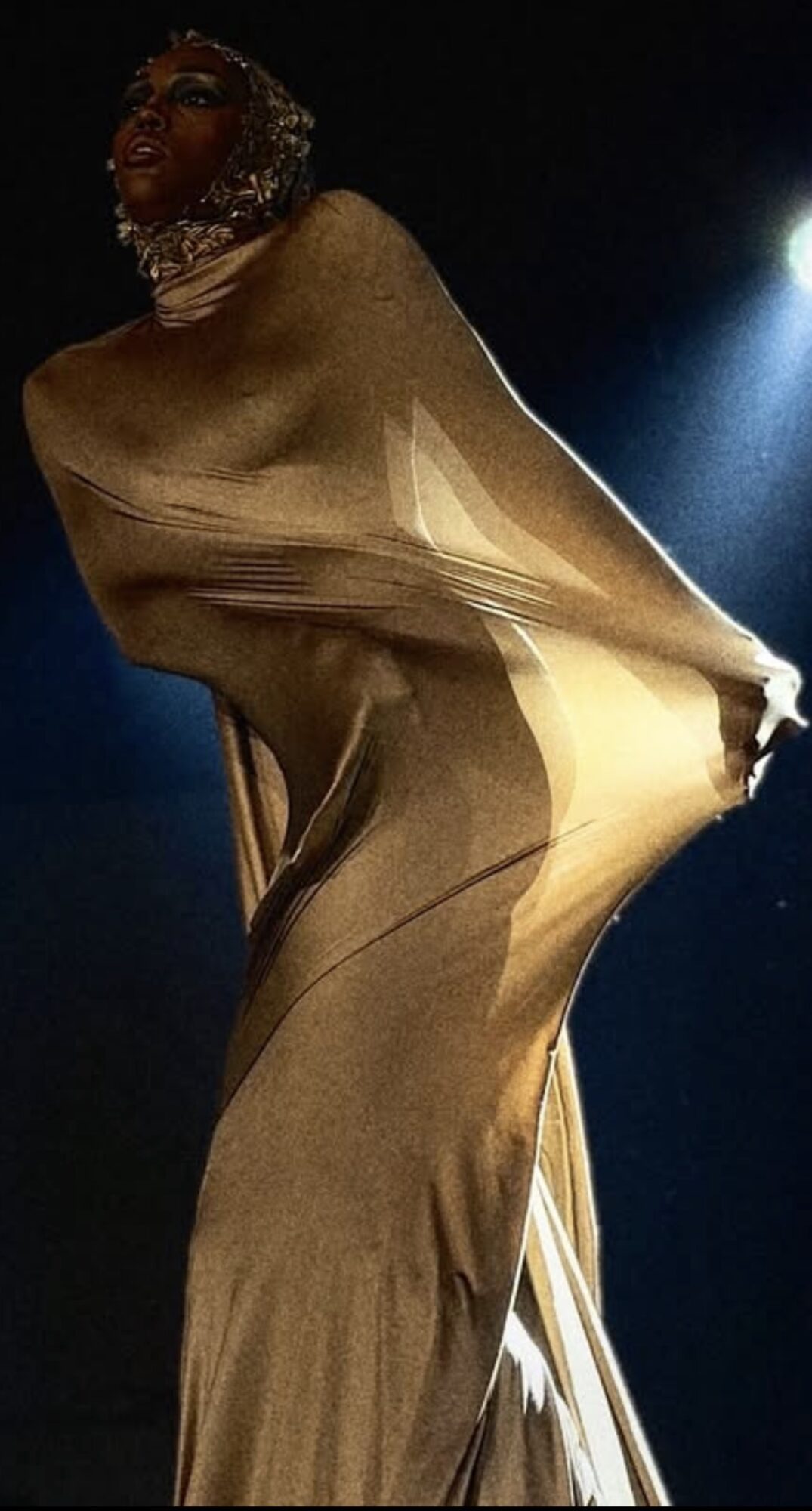 Person in a shiny gold dress on stage, holding the dress out to the side, with a dark background and bright light.