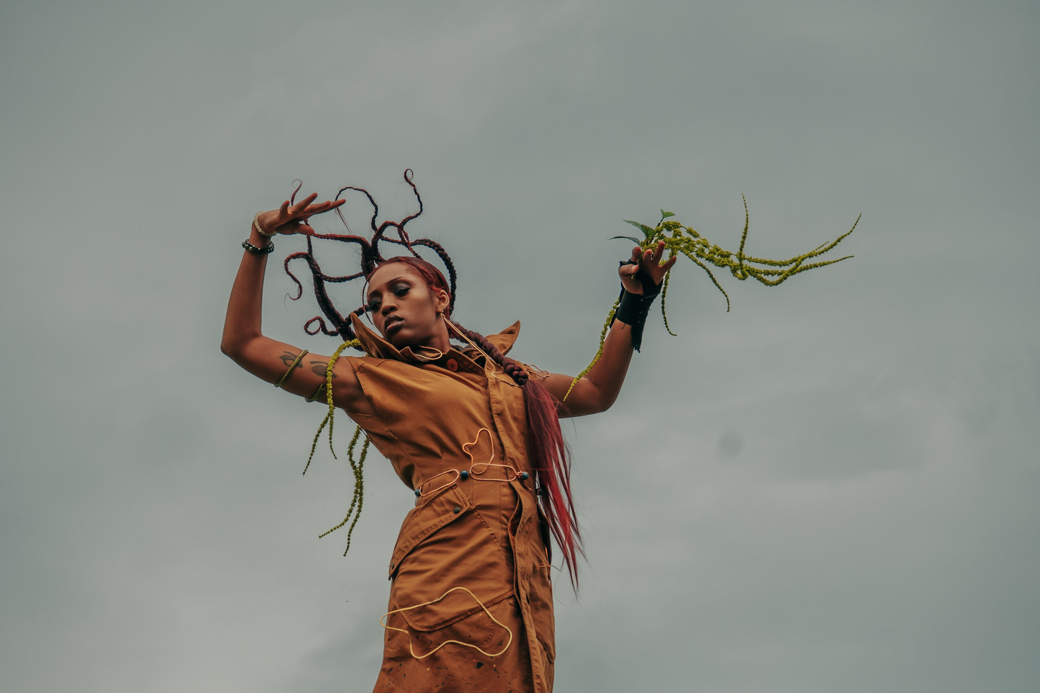 A person with dreadlocks and colorful hair stands against a cloudy sky, holding plants in both hands.