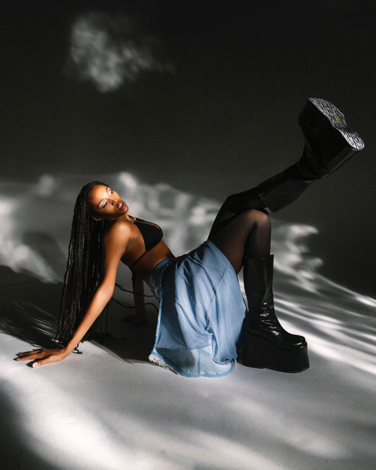 Woman with long hair lying on a reflective surface, wearing black top, denim skirt, and black boots, with a dark background.