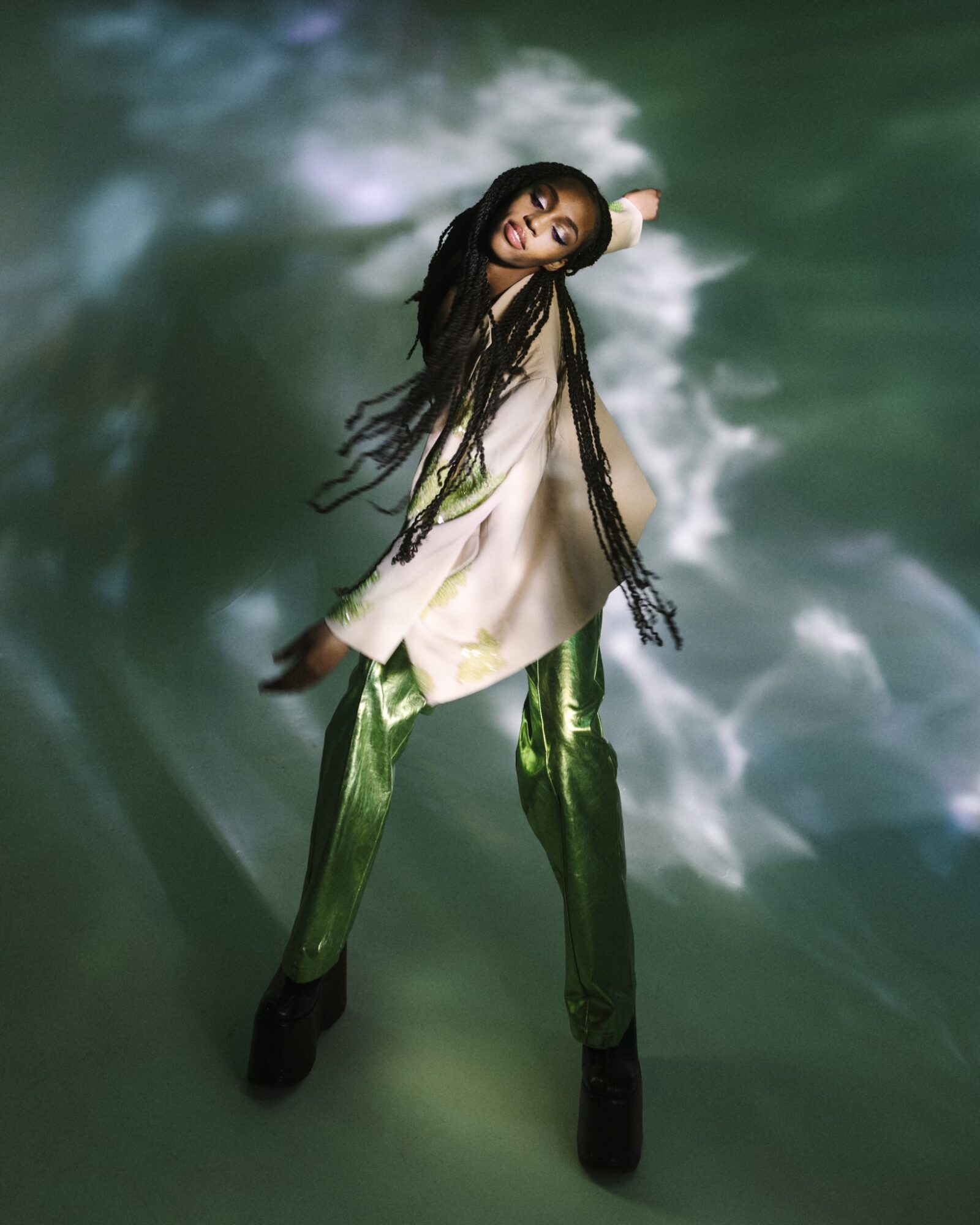 Woman with long hair poses against a cloudy sky background, wearing a white top and shiny green pants.