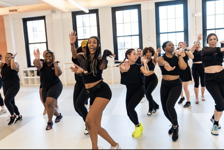 Group of women dancing in a bright studio with large windows, smiling and moving energetically.