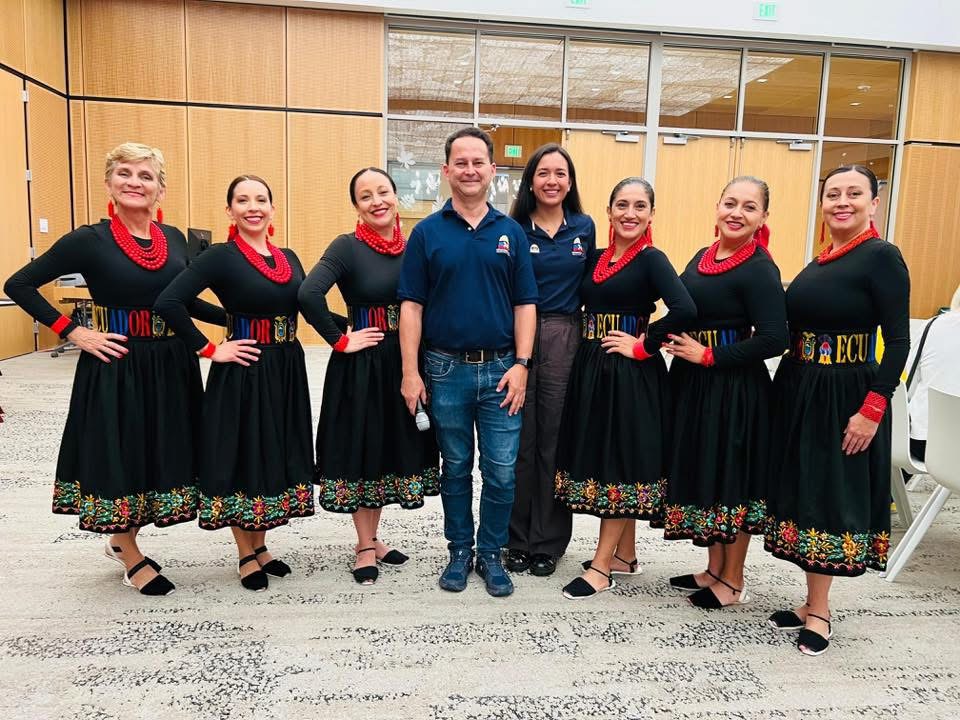 Seven women in traditional dresses and two men in casual clothing stand together indoors, smiling, with wooden paneling and glass windows behind them.