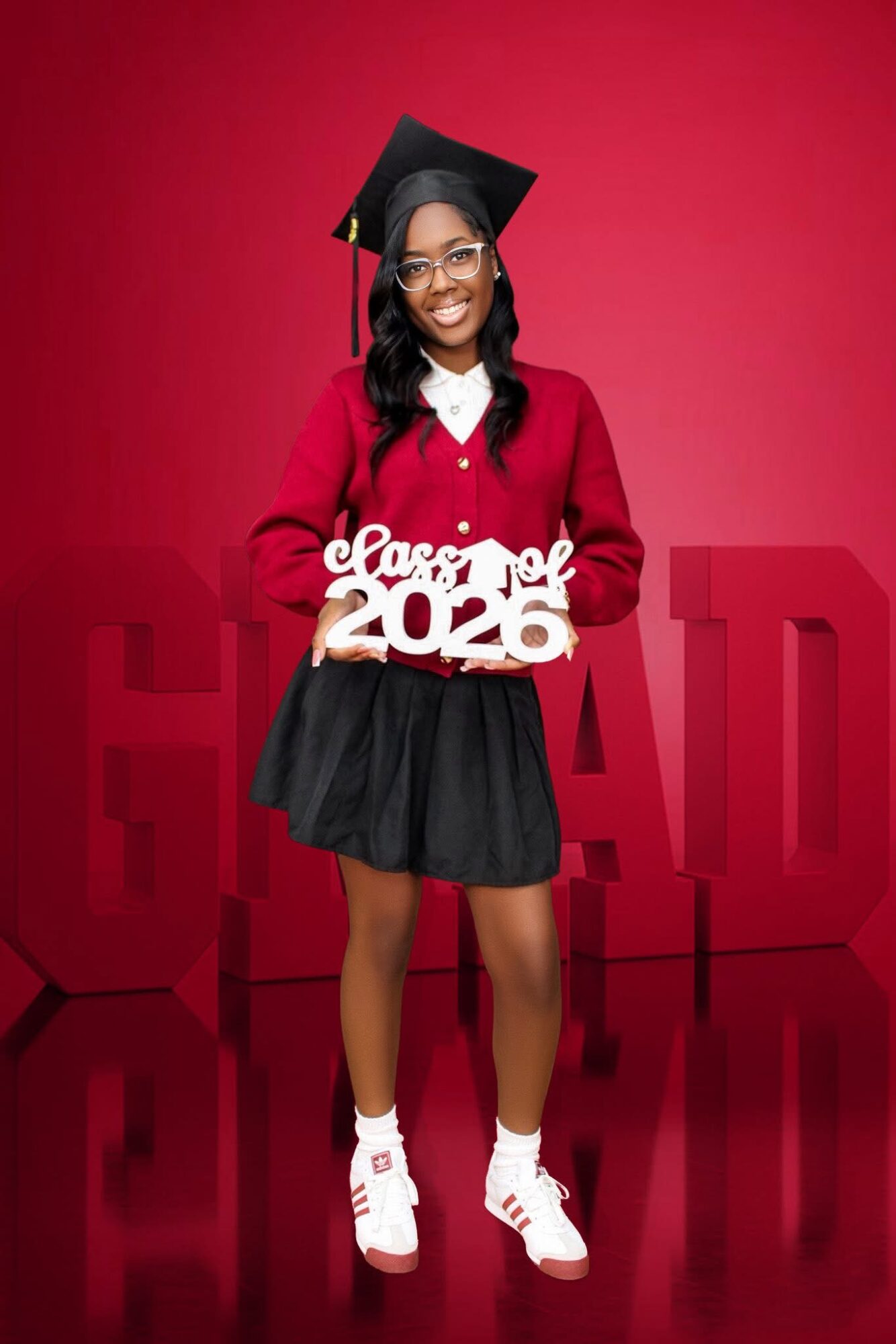 Young woman in graduation cap and gown holding a sign that says 'Class of 2026' against a red background.