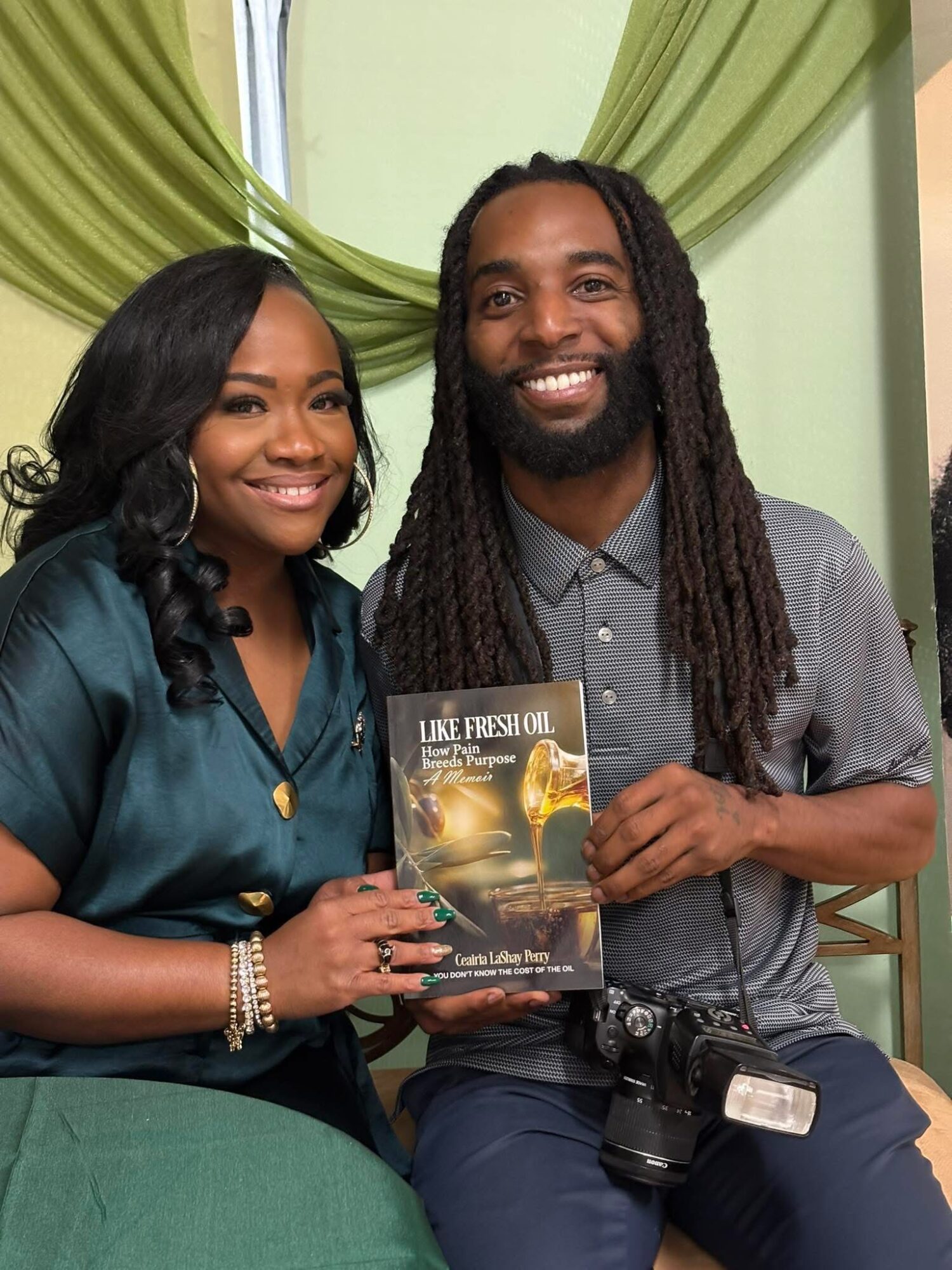 Two smiling people holding a book and a camera, sitting in front of green curtains.