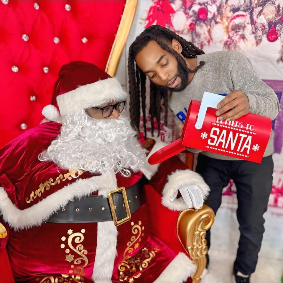 Person dressed as Santa Claus sitting on a red and gold chair, talking to a man holding a gift box, Christmas decorations in background.