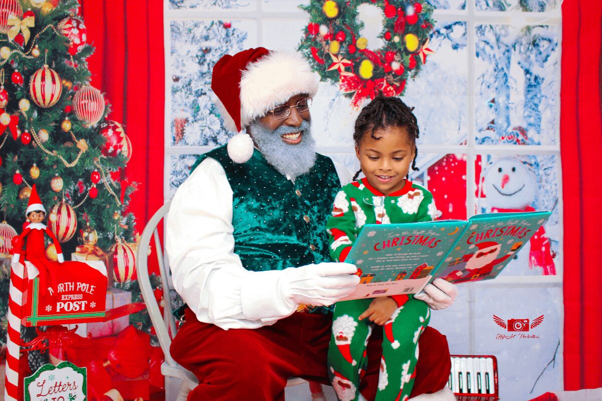 Man dressed as Santa Claus reading a Christmas book to a child, with Christmas decorations and a tree in the background.