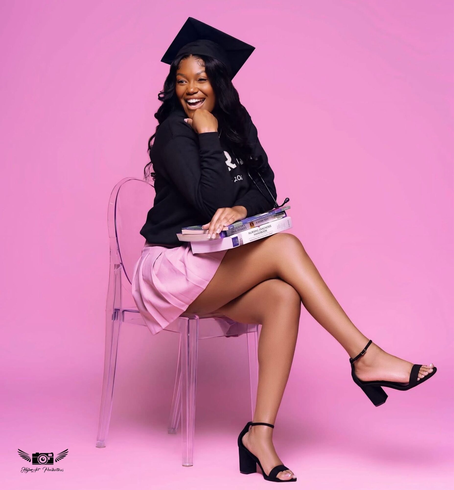 Young woman in graduation cap and gown, sitting on a transparent chair, smiling, holding a diploma, against pink background.