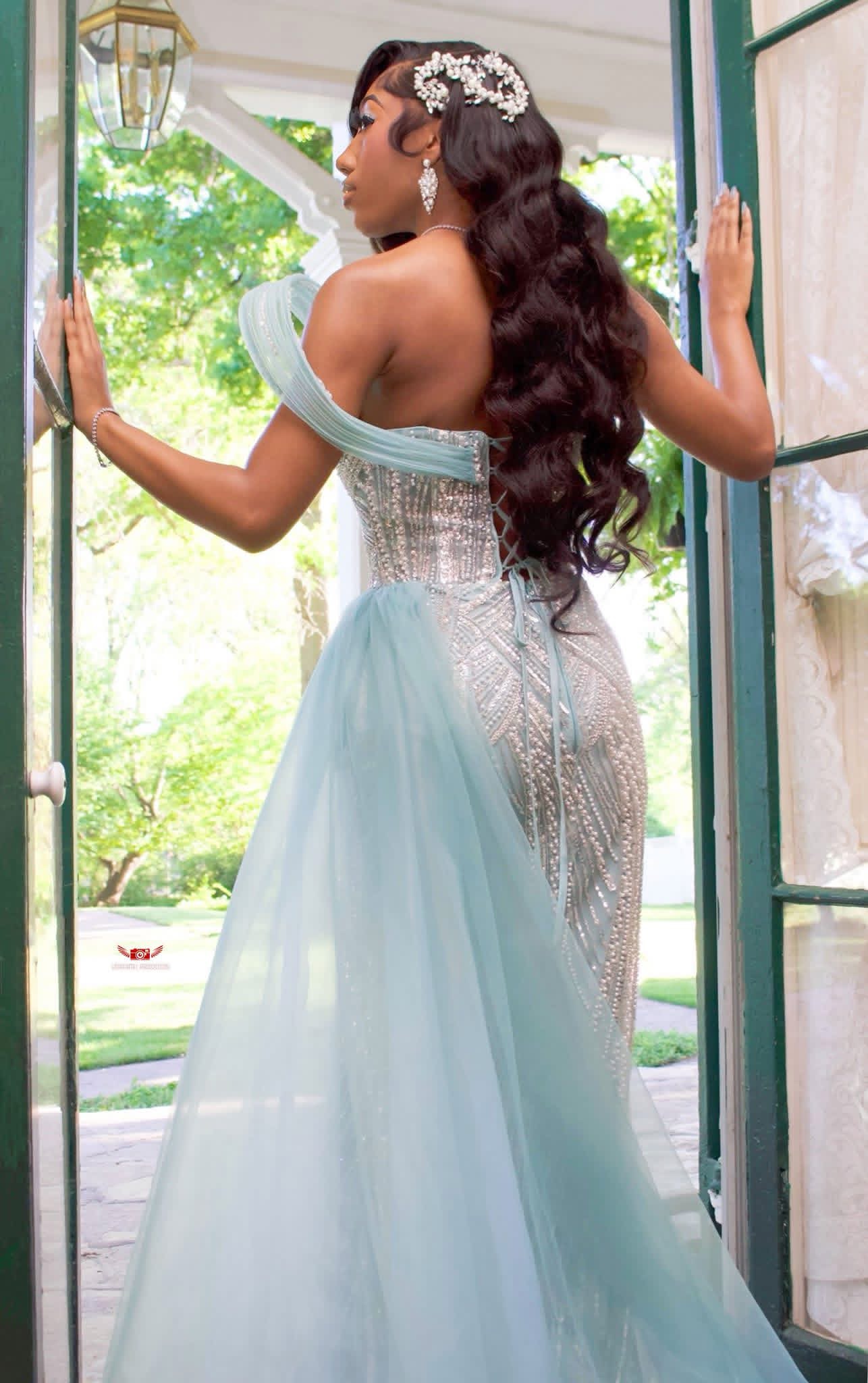 Woman in a light blue gown standing at an open door, looking outside with her back to the camera.