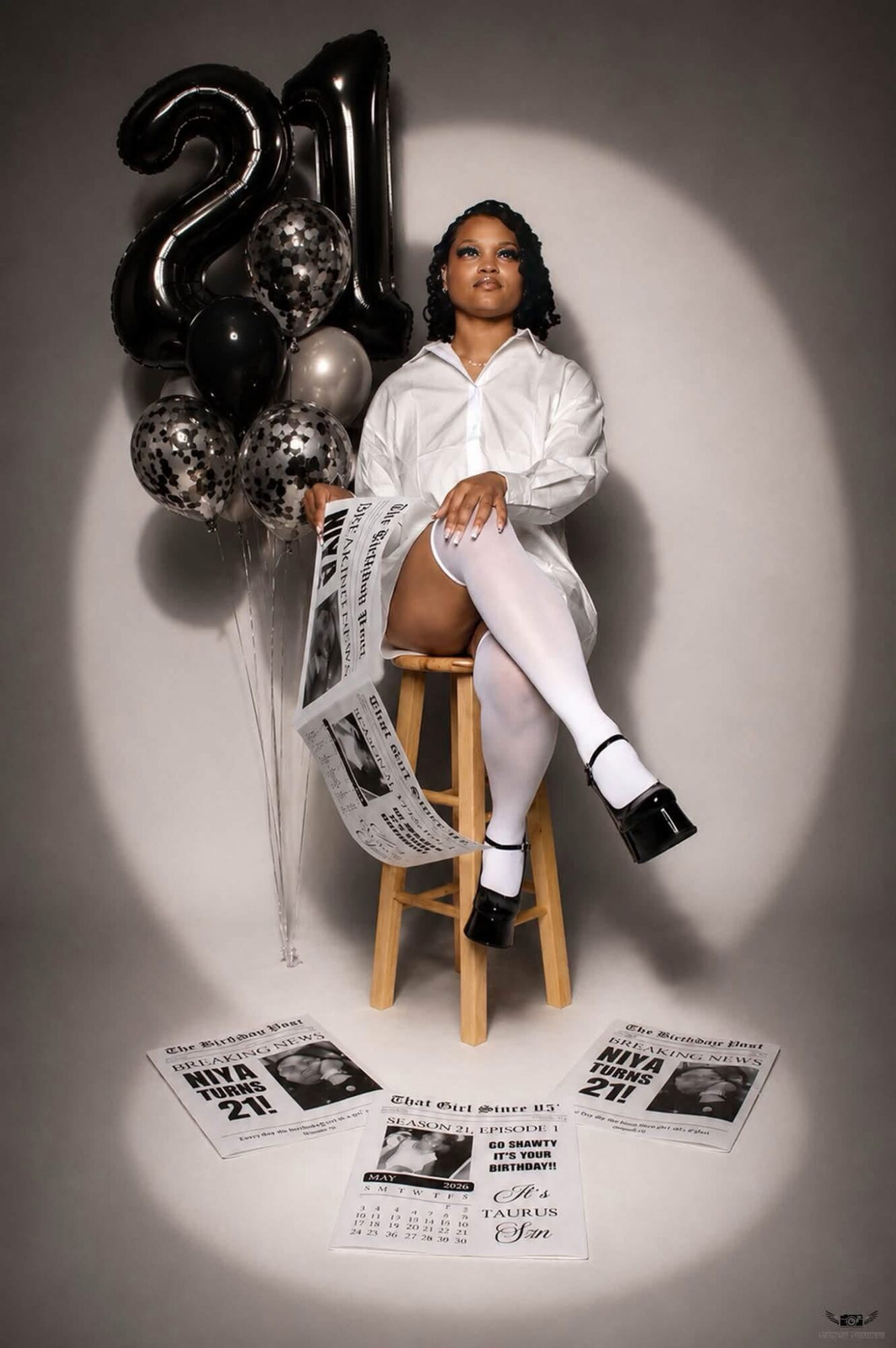 Woman sitting on a stool with black and silver balloons shaped as '21' behind her, holding newspapers, in a spotlighted setting.