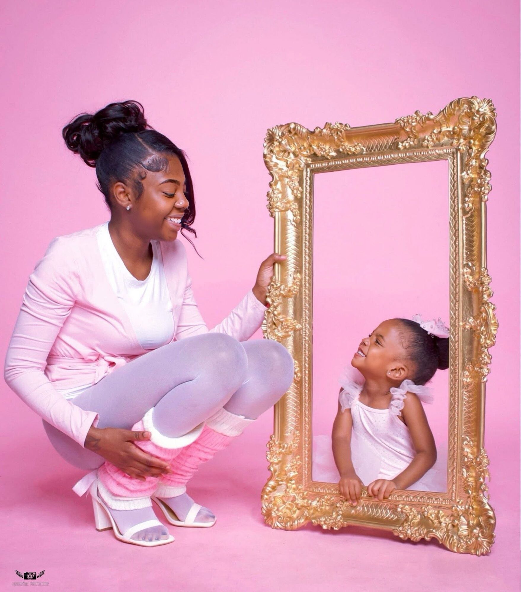 Woman and girl looking at each other through a large ornate gold frame against pink background.