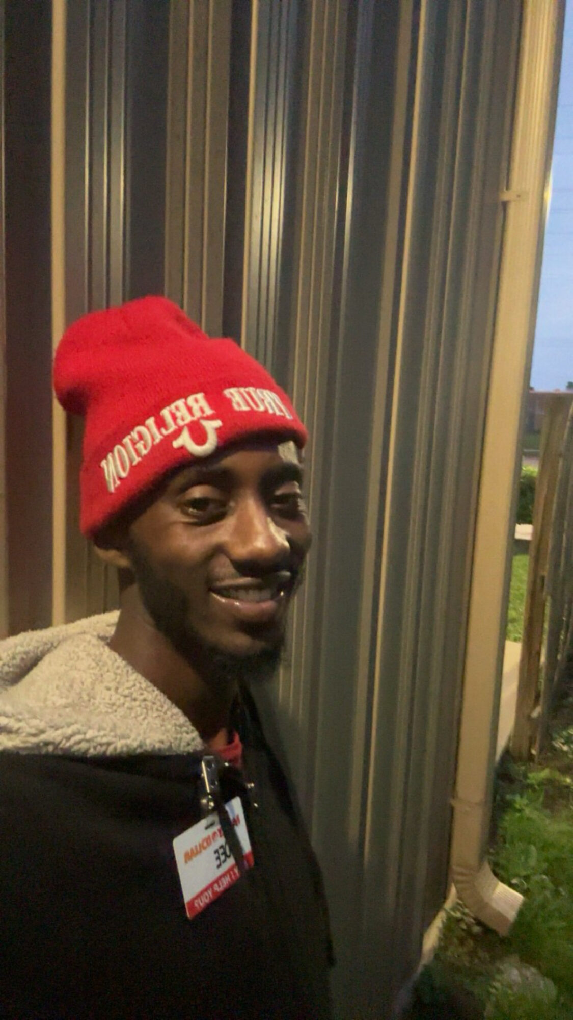 Person wearing a red beanie and black jacket, standing outdoors near a metallic wall, smiling.