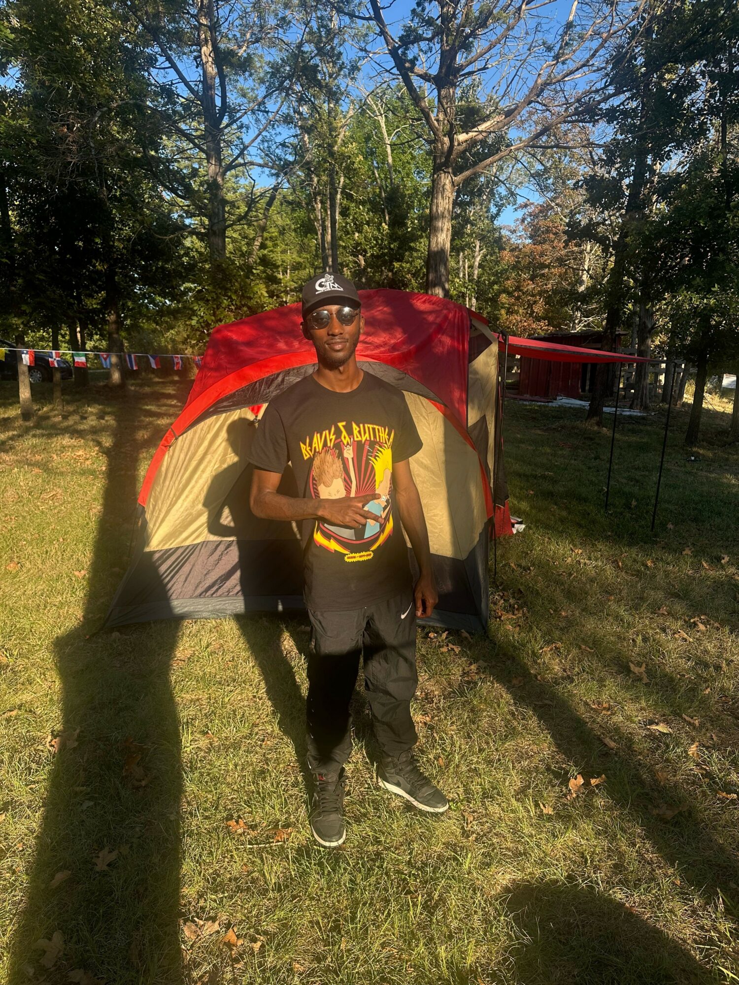 Person standing outdoors in front of a tent with trees and grass, holding a drink, wearing a Batman shirt and cap.