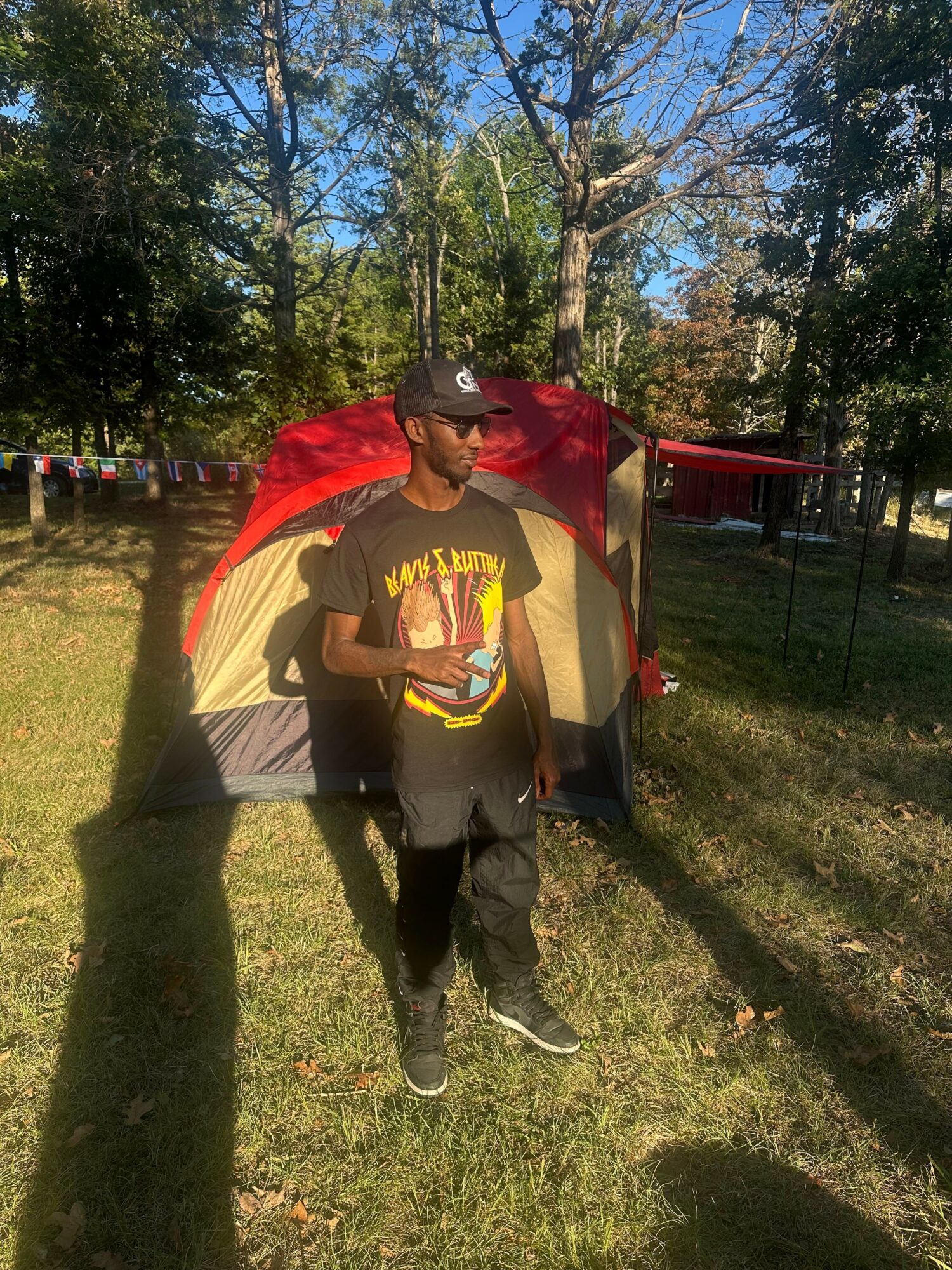 Person standing outdoors near a tent with trees in the background, holding a phone, wearing a black cap and T-shirt.