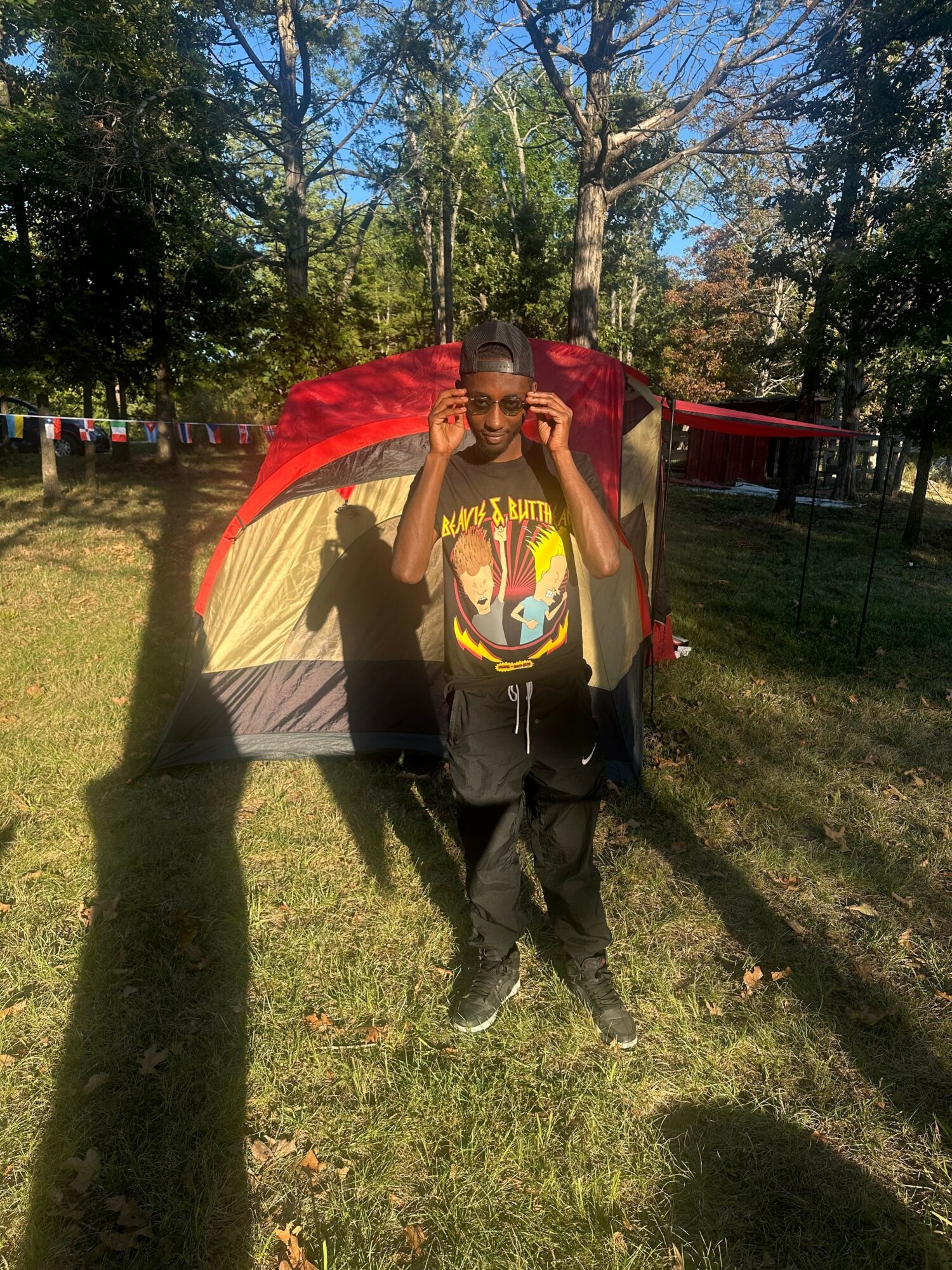 Person standing outdoors near a tent, holding a phone, with trees and grass in the background.
