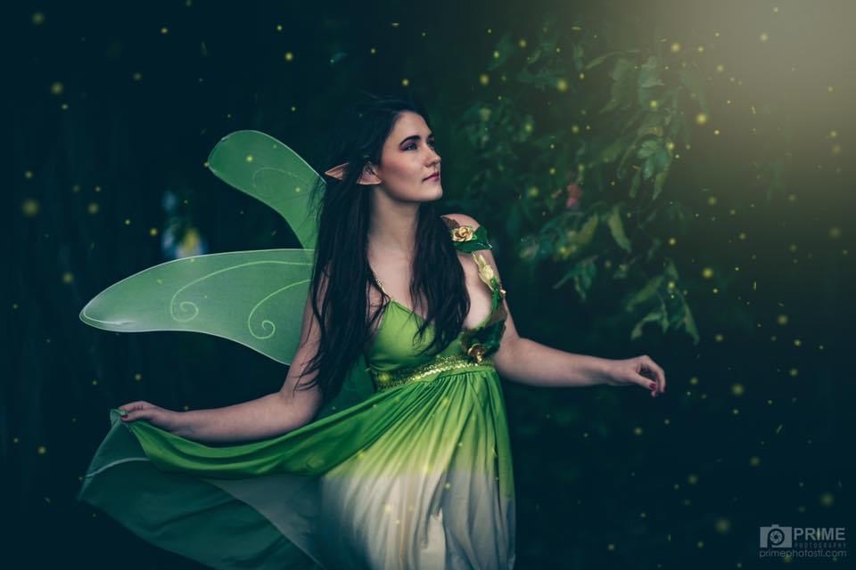 Woman dressed as a fairy with green wings and dress, standing outdoors with a dark background and light effects.