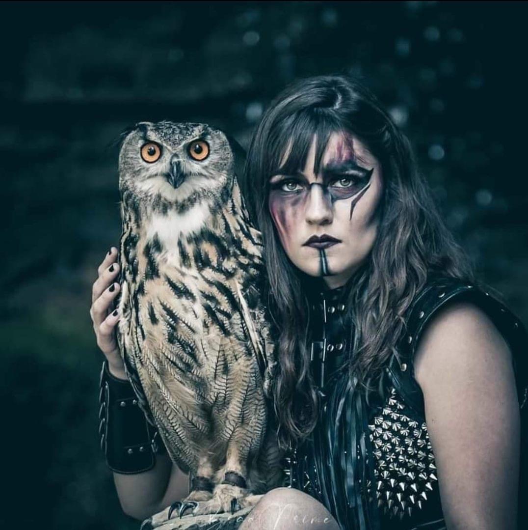 Person with dramatic makeup holding an owl, dark background, wearing black clothing with spikes.