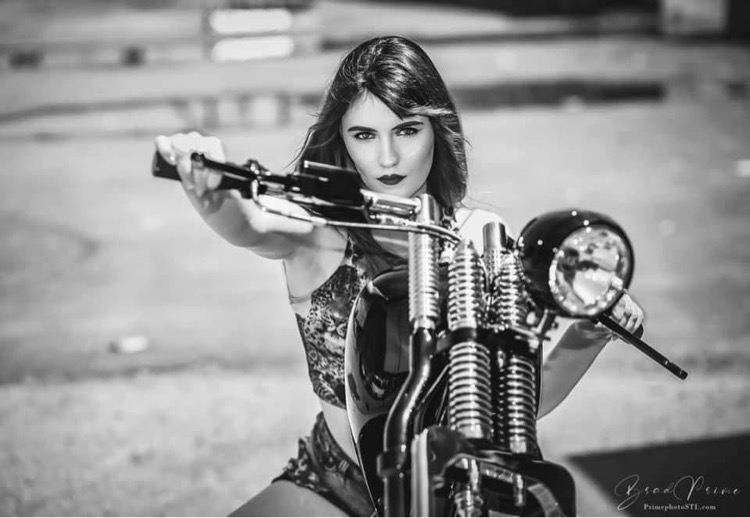 Woman with long hair sitting on a motorcycle, holding the handlebars, looking at the camera, outdoors, black and white.