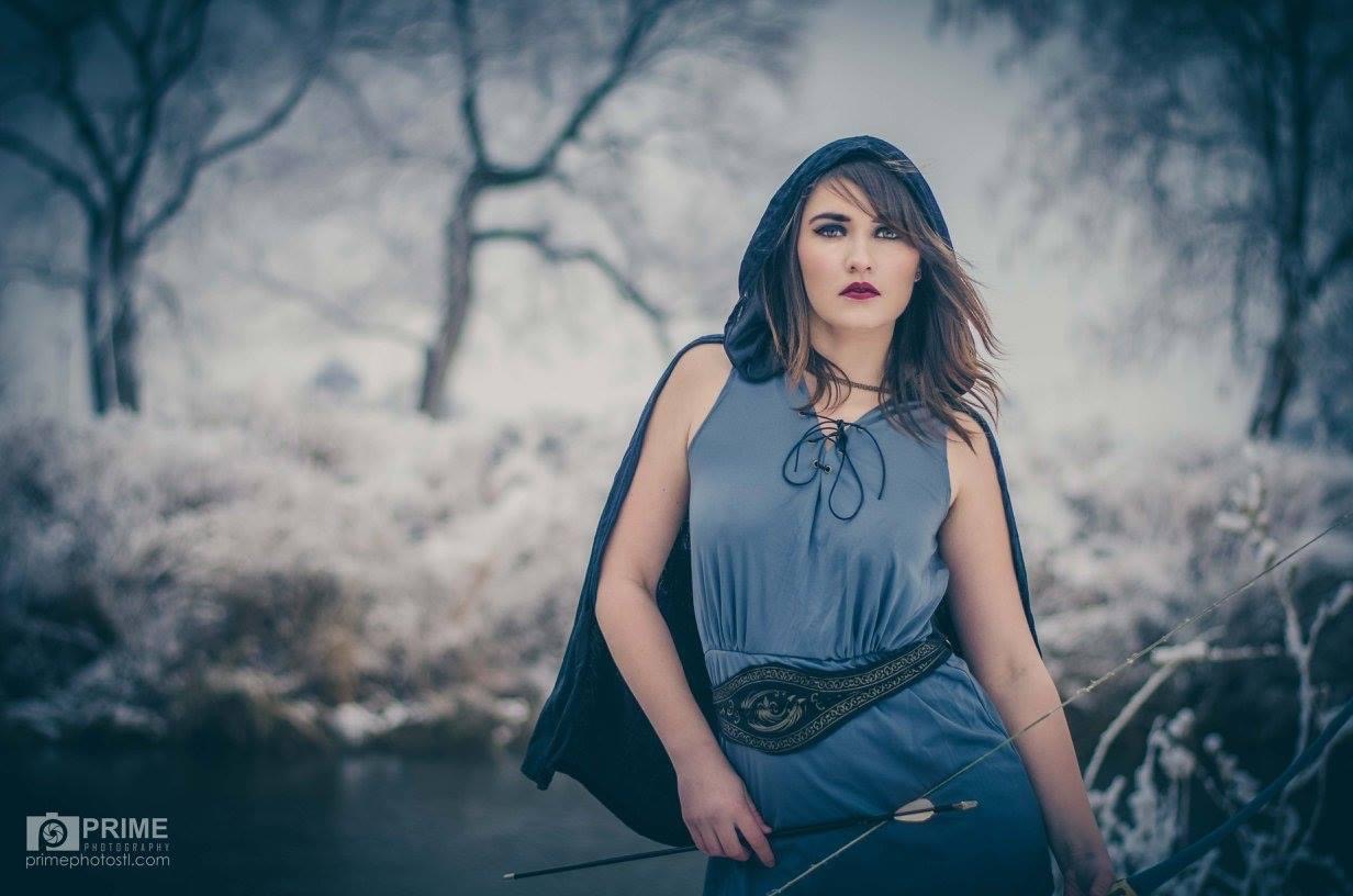 Woman with dark hair and makeup in a blue dress outdoors in a snowy landscape, trees in background.