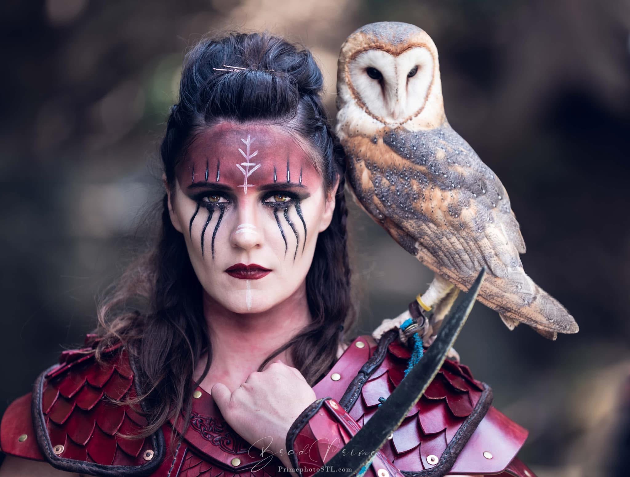 Woman with face paint and costume holding an owl on her shoulder, outdoors, dark background.