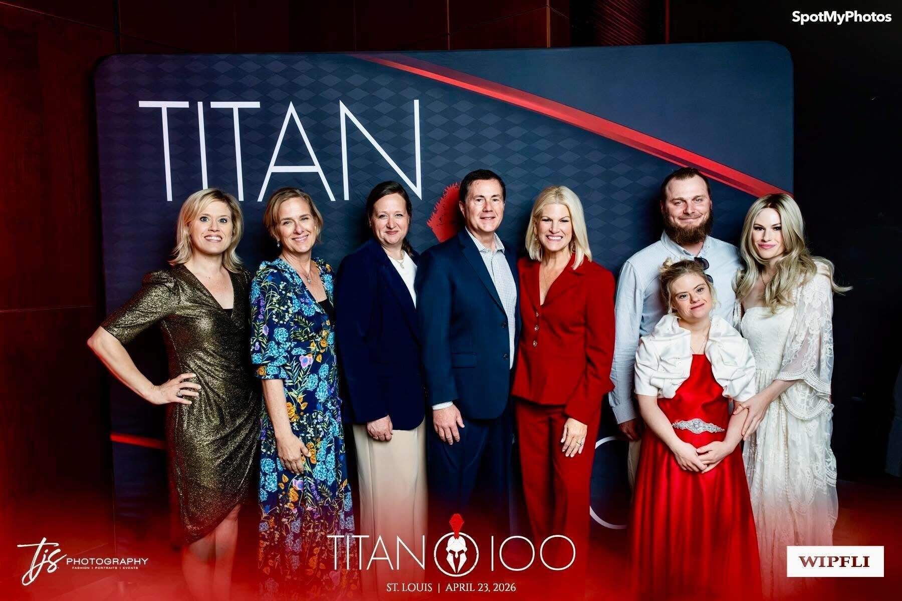 Group of eight people standing in front of a Titan 100 backdrop, posing for a photo, some smiling, some serious.