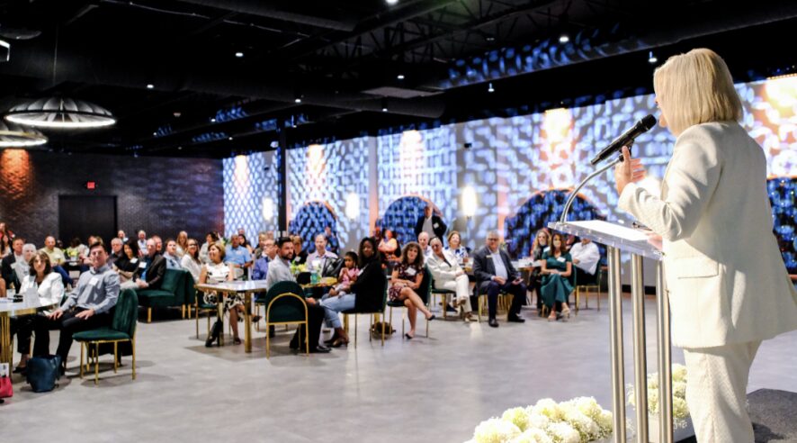A woman in a white suit speaks at a podium to an audience in a large conference room with blue patterned windows.