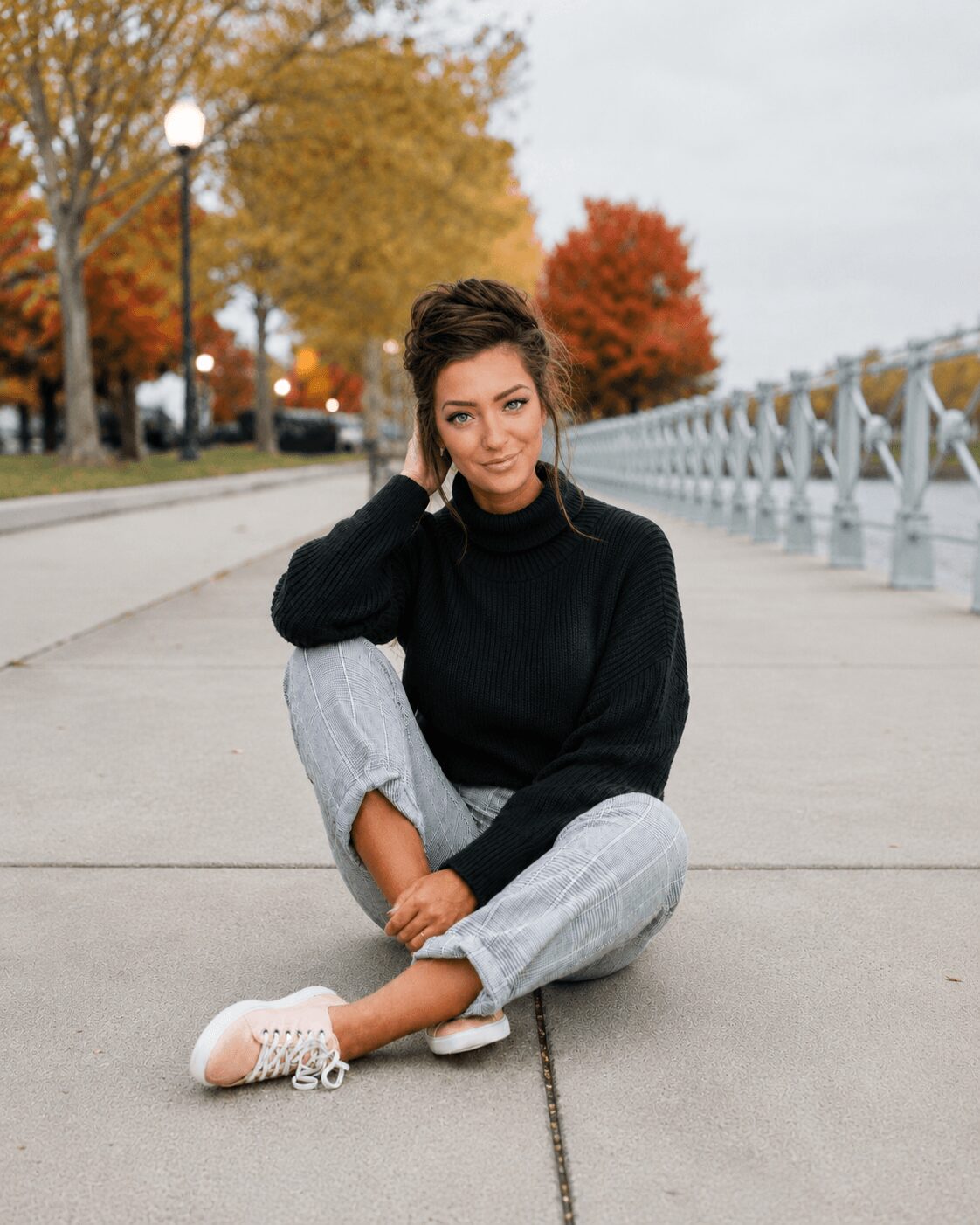 Woman sitting on sidewalk with autumn trees and railing in background, smiling, wearing black sweater and light pants.