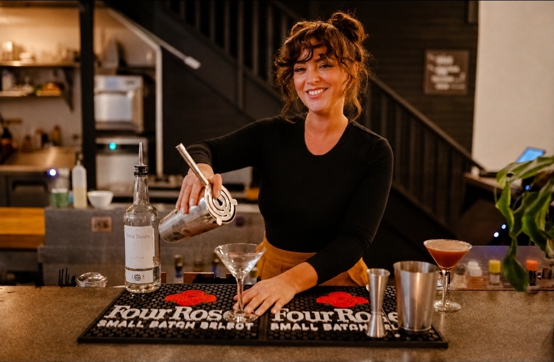 Woman smiling behind bar counter with drinks and bar tools, warm indoor setting, plants, and kitchen in background.
