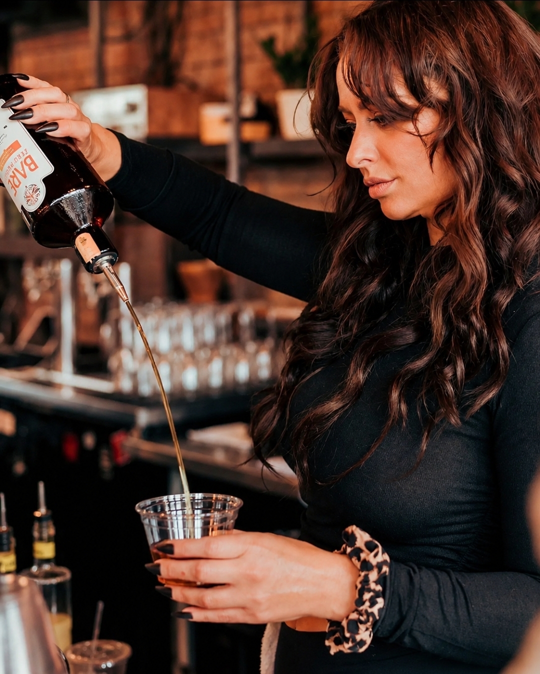 Woman pouring drink into a glass in a bar setting, with shelves and glasses in the background.