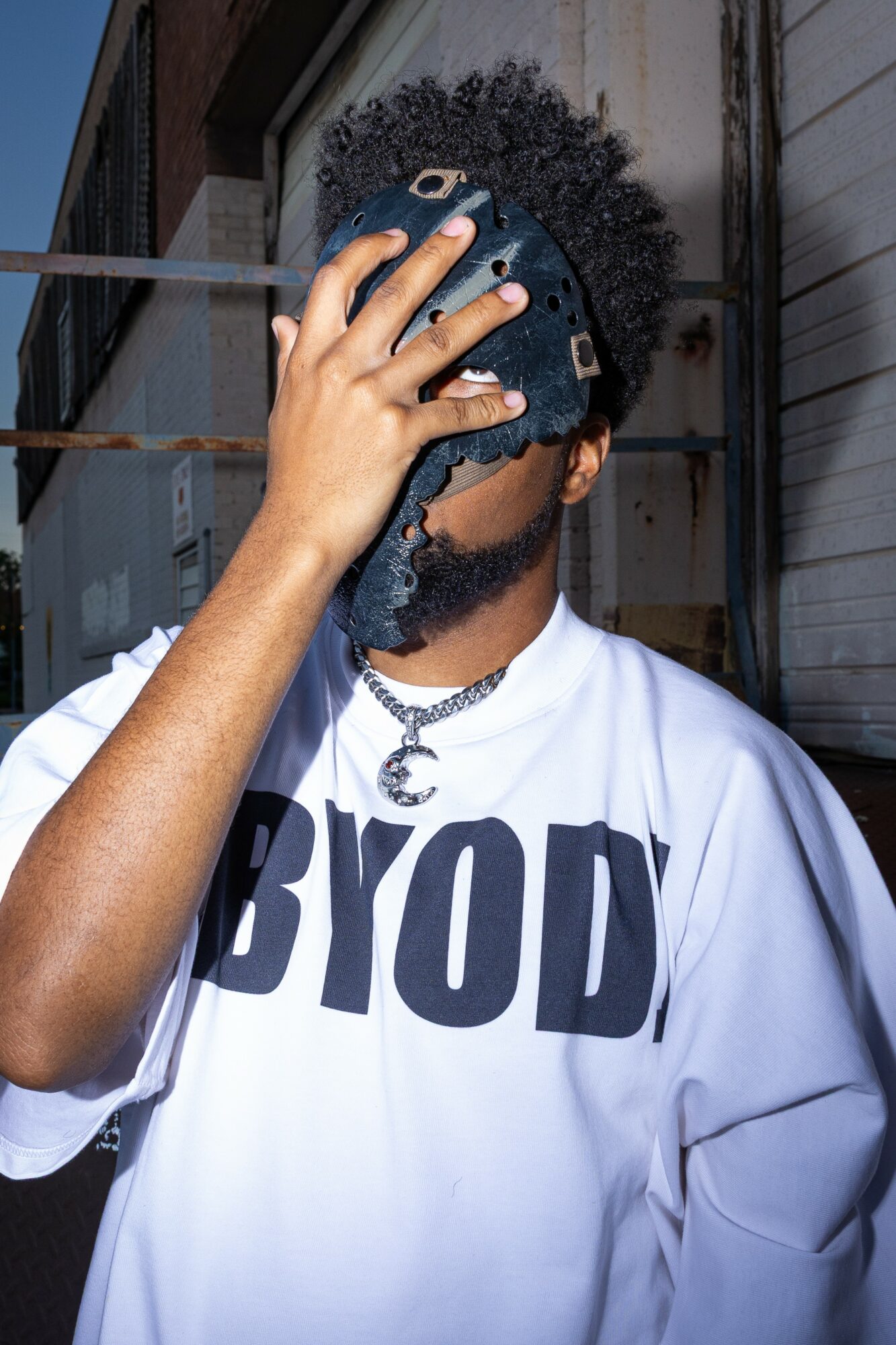 Person with curly hair wearing a white BYOD shirt, holding a mask over their face, standing outdoors.