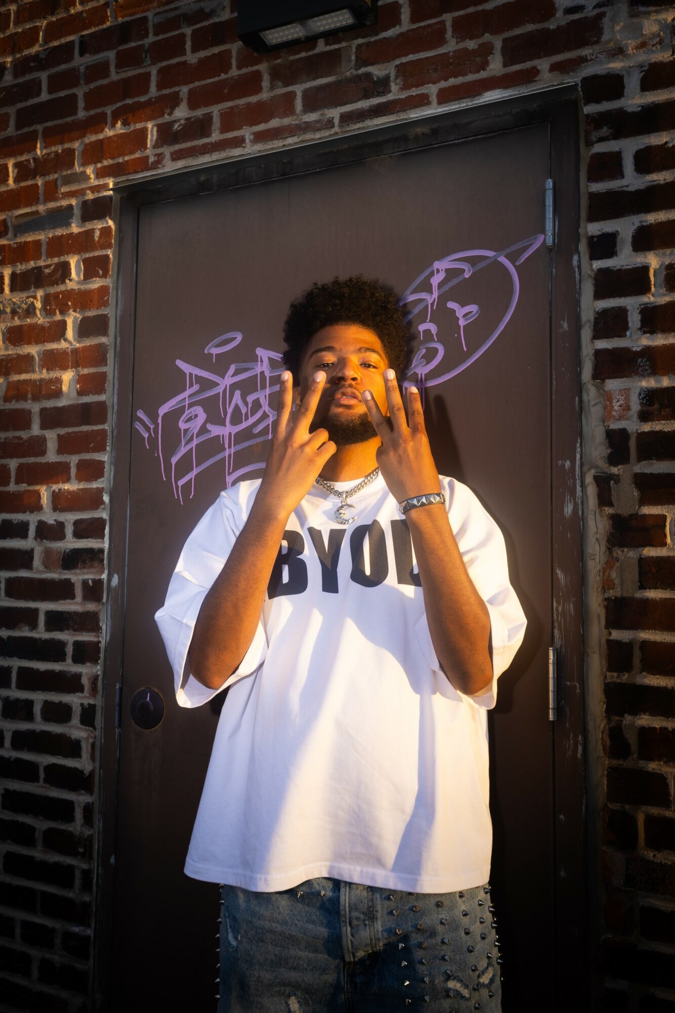 Young man with curly hair and beard standing in front of a black door with purple graffiti, wearing a white T-shirt and jeans.