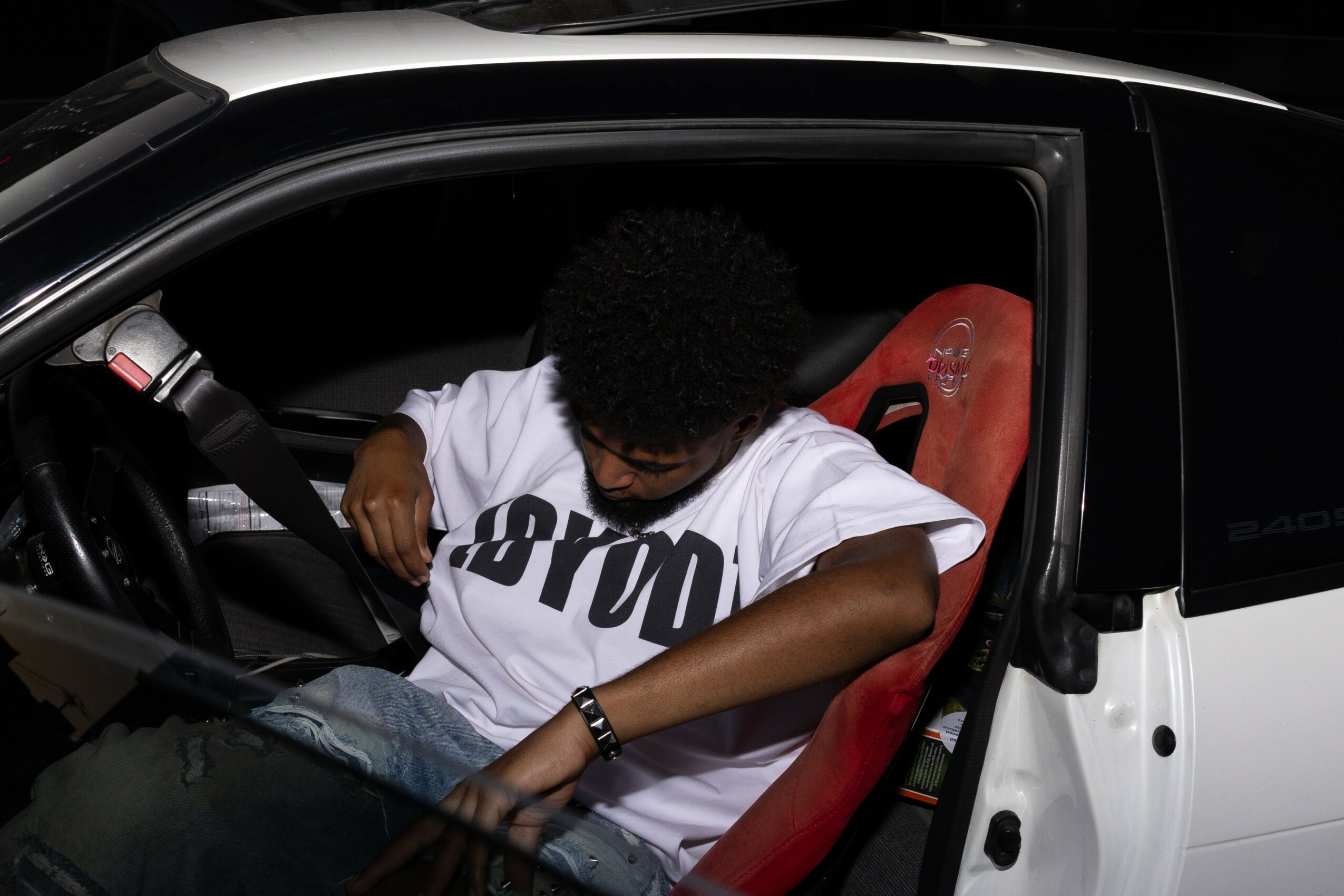 Person with curly hair sitting in a car seat, wearing a white T-shirt, with a dark background