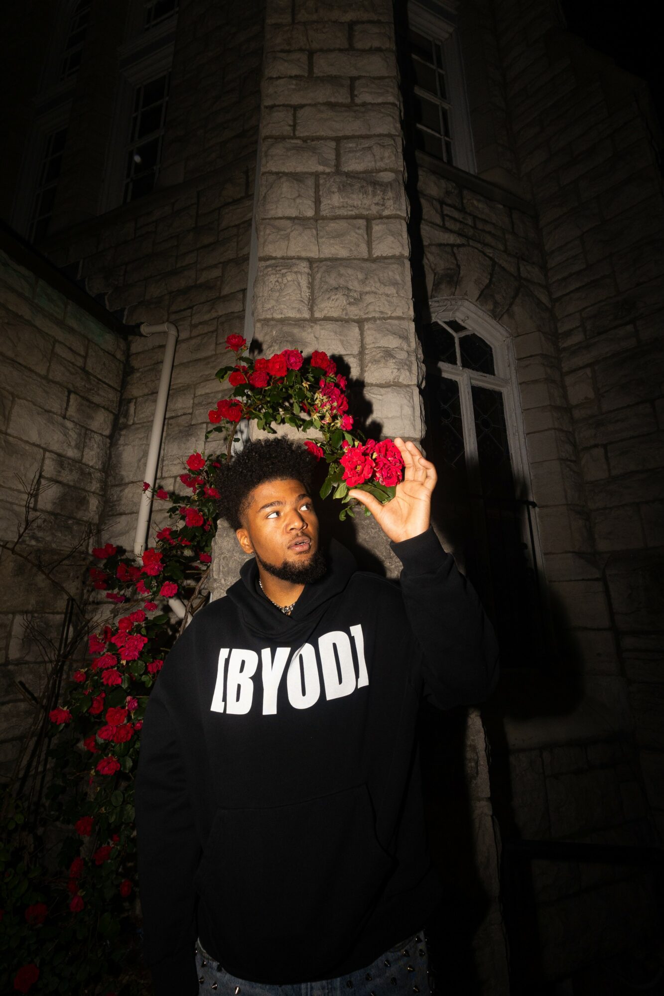 Young man with a hoodie standing in front of a stone building and red flowers at night.