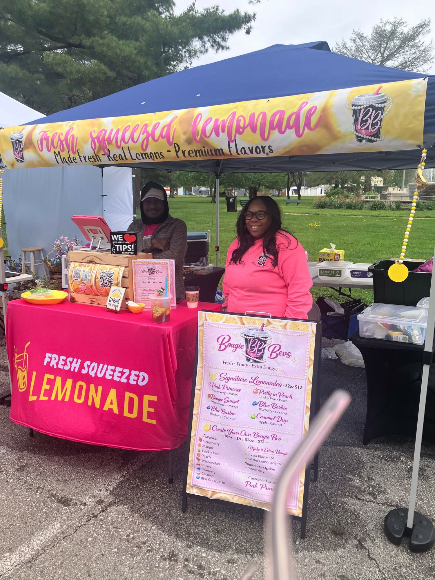 Two women stand behind a table with a lemonade stand sign, outdoors under a blue canopy, with trees in the background.