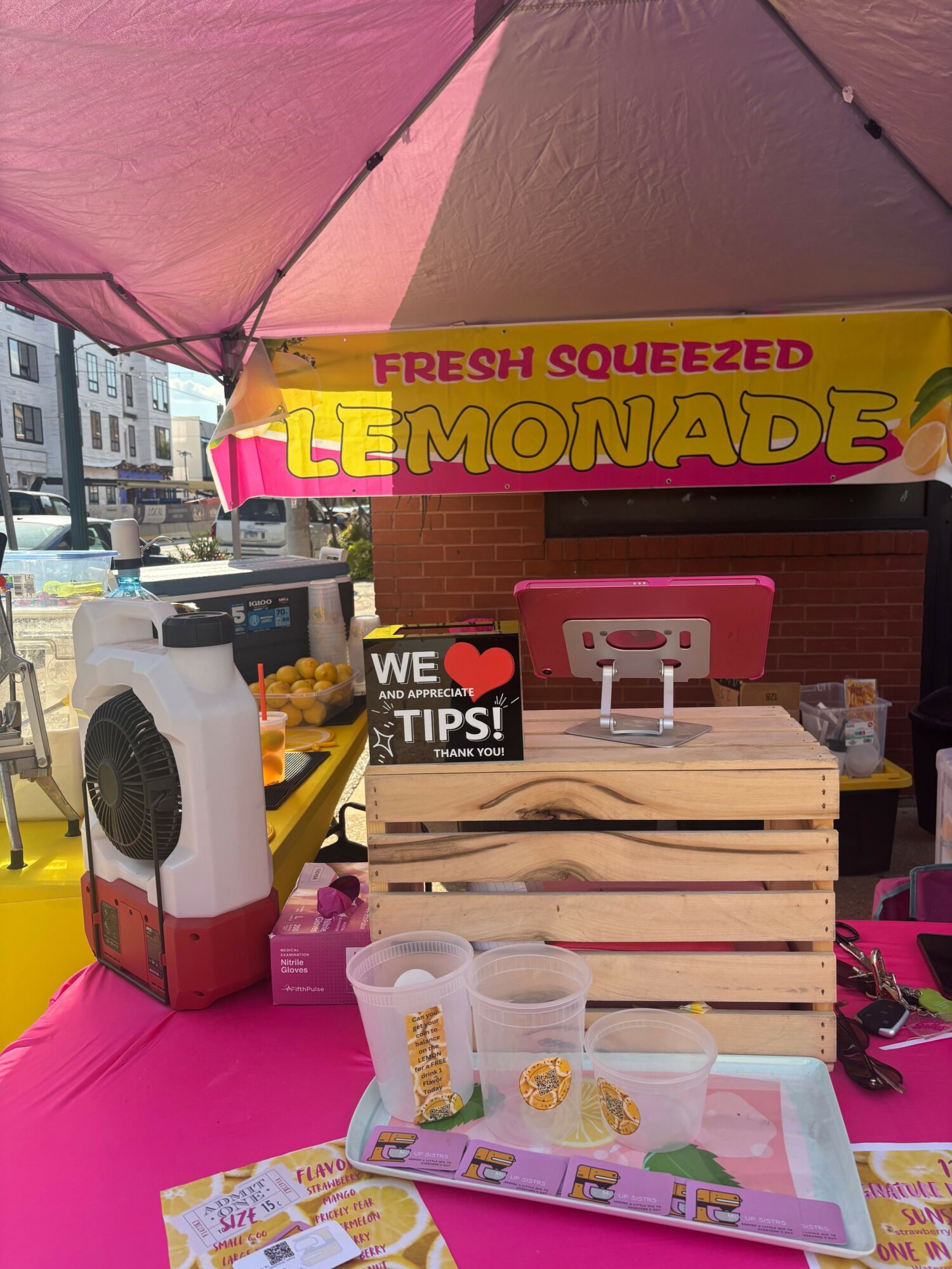 Display with lemonade sign, wooden crate, and various items on pink surface, viewed from the side.