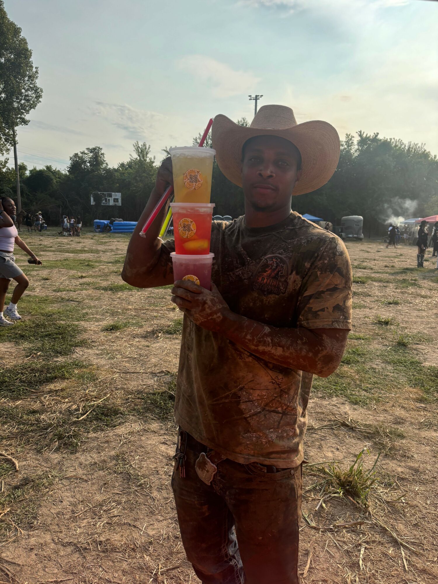 Person wearing a hat holding a box outdoors on a dirt field with trees and cloudy sky in background.