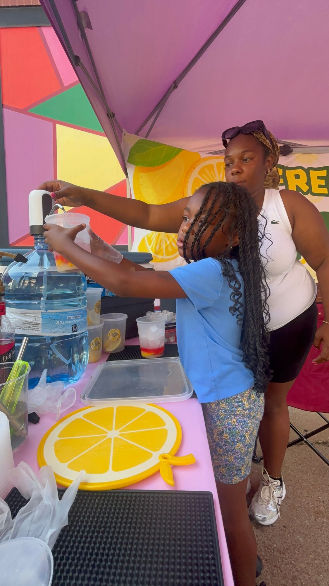 Two girls at a colorful outdoor stand, one pouring water into a container, with a lemon-shaped tray nearby.