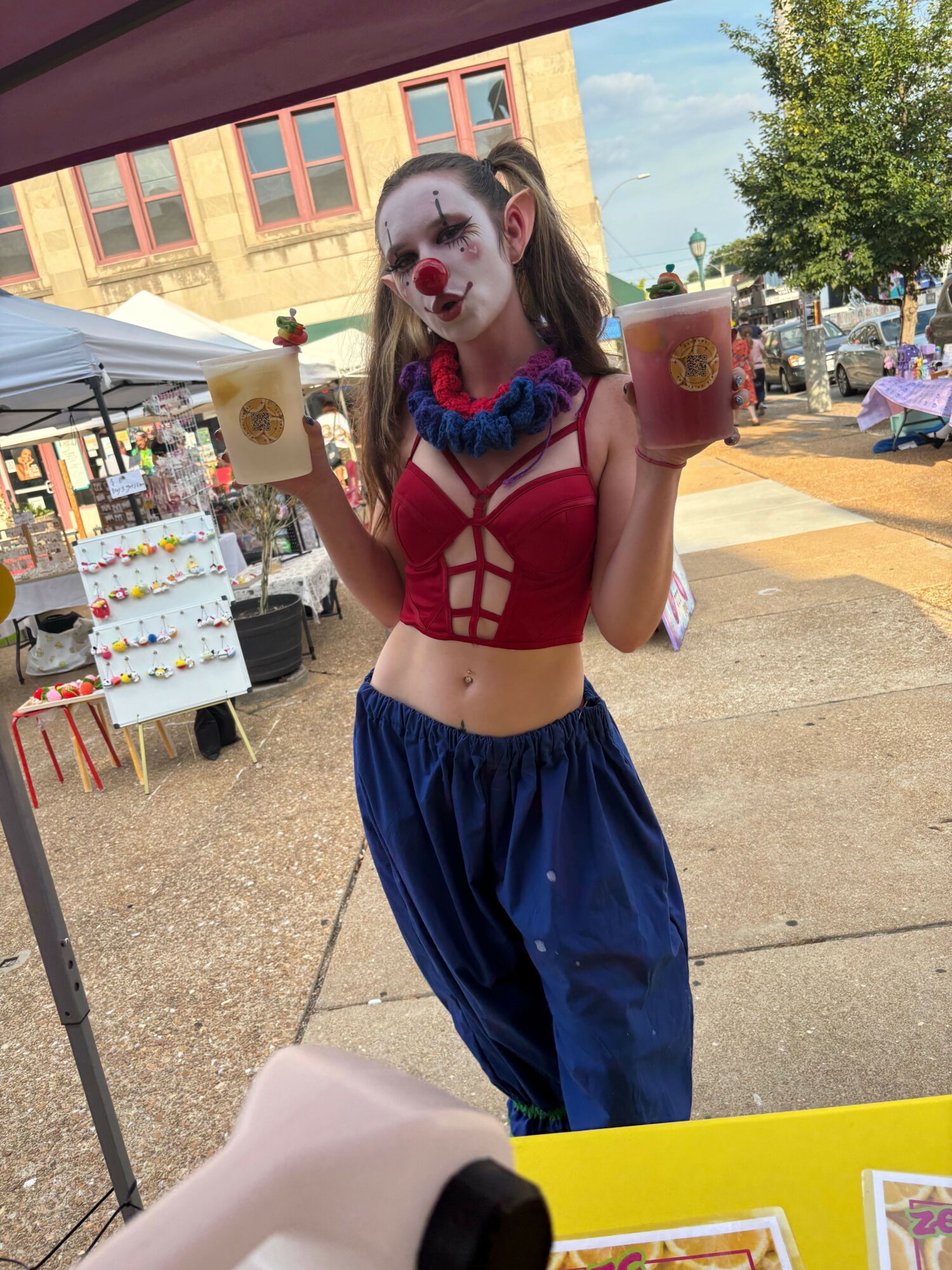 Person dressed as a clown with face paint, red and blue costume, standing outdoors near a table and building.