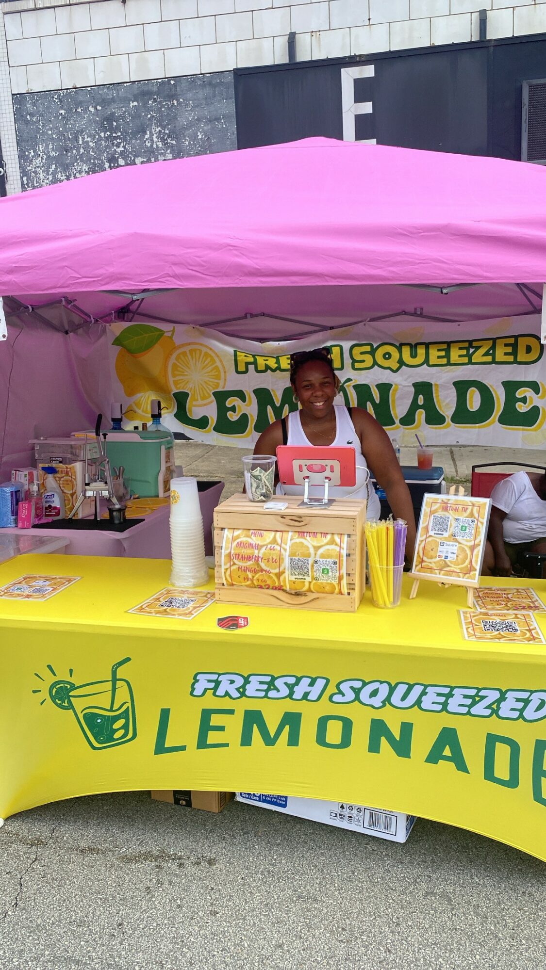 Person standing behind a lemonade stand with a pink canopy, yellow tablecloth, and lemon decorations, outdoors.
