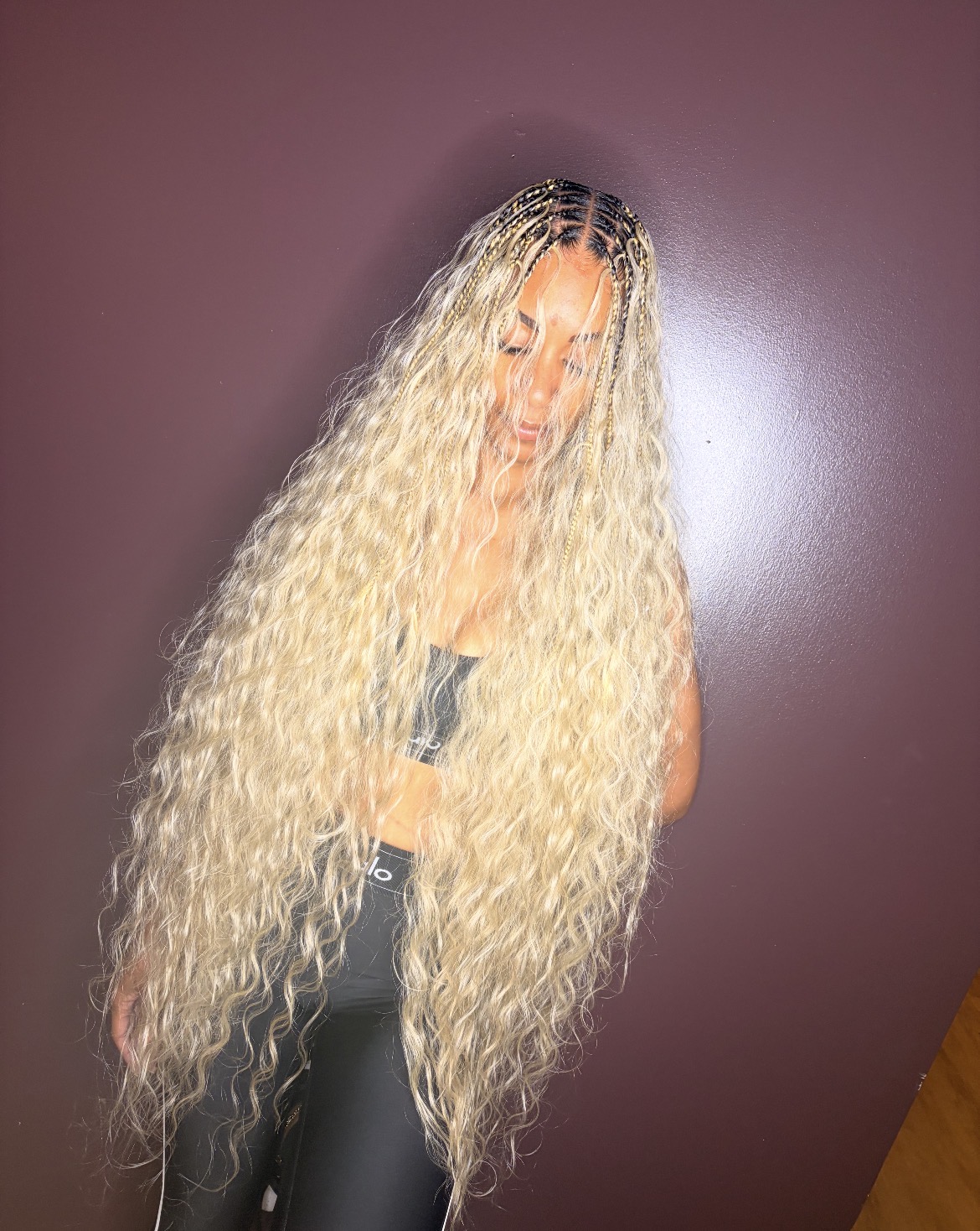 Person with long, curly blonde hair standing against a maroon wall, partially covering face with hand.