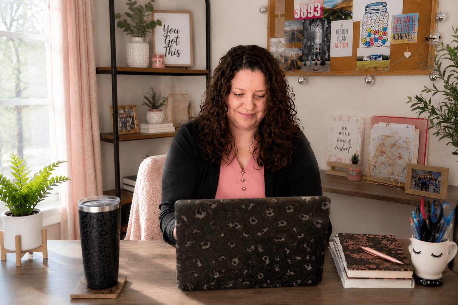 Woman with curly hair working on a laptop at a desk in a cozy room with plants and shelves.