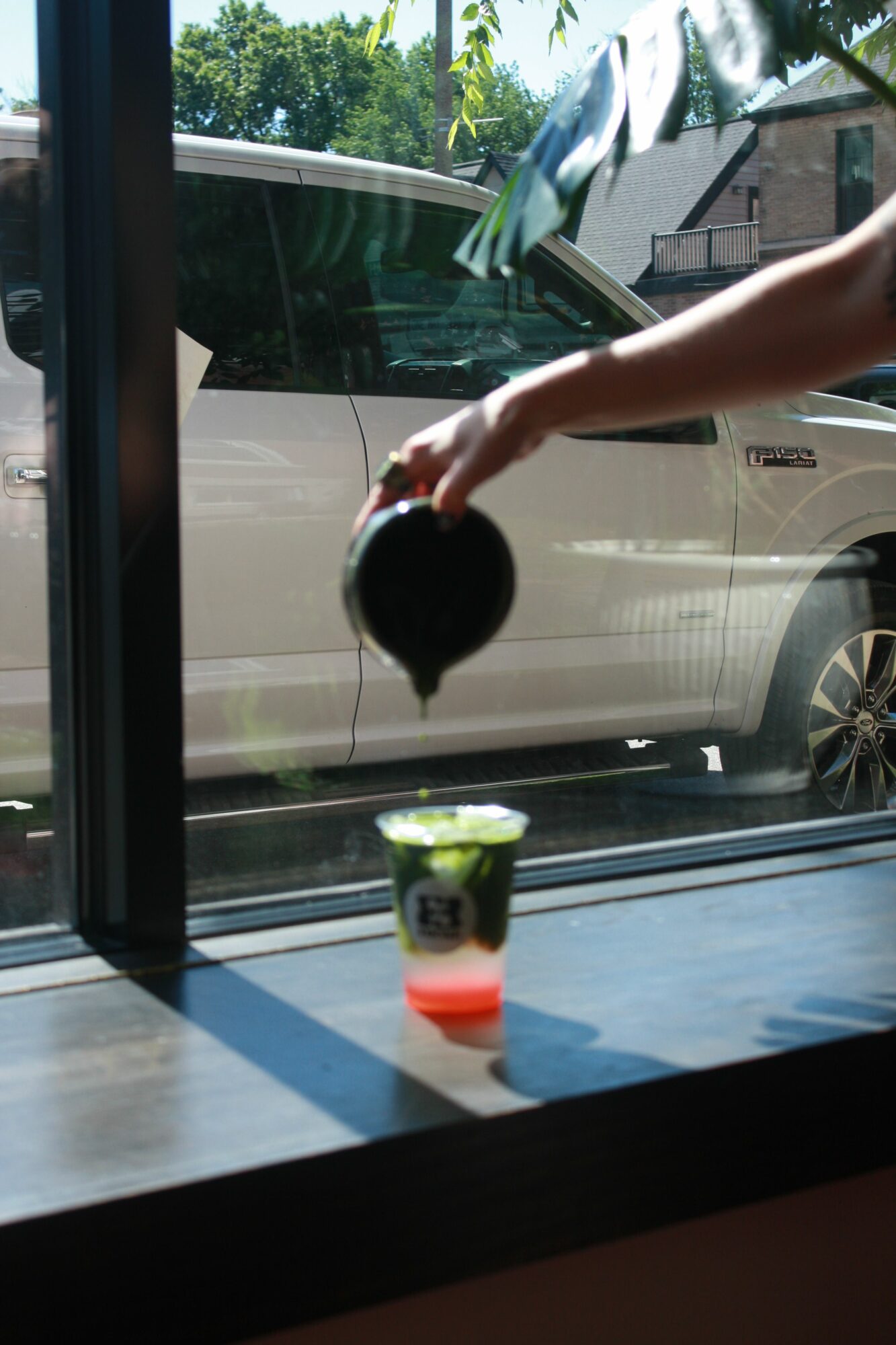 Person pouring a dark liquid from a small container into a glass on a windowsill, with a car outside.
