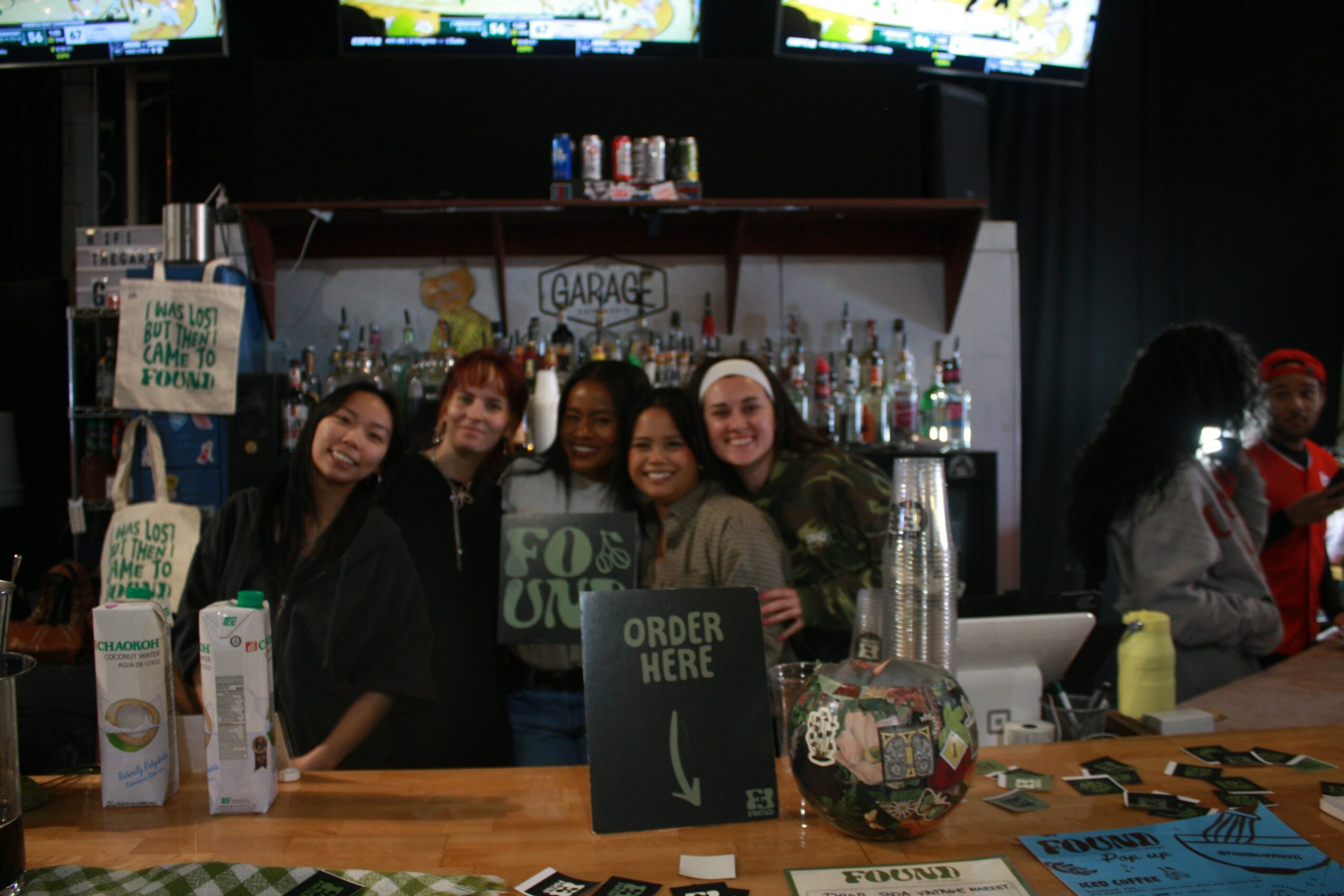 Five people standing behind a bar counter smiling, with bottles and signs, in a dimly lit setting.