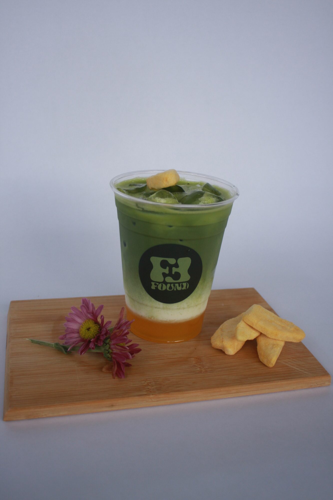 A green and orange drink in a clear cup with a black logo, on a wooden tray with snacks and a flower.