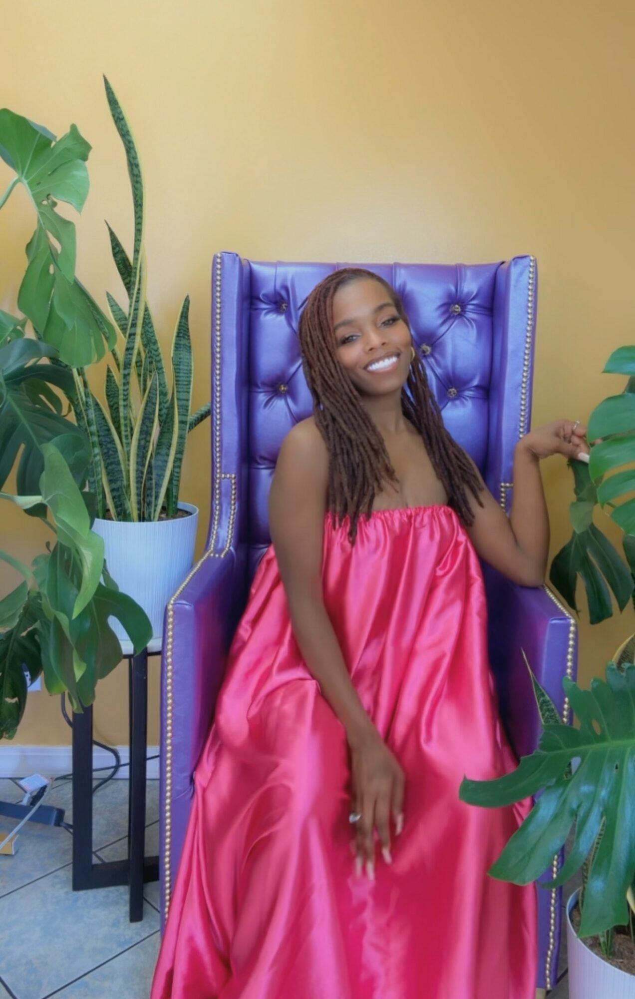 Young woman with long hair sitting on a purple chair, smiling, wearing a pink dress, surrounded by green plants.