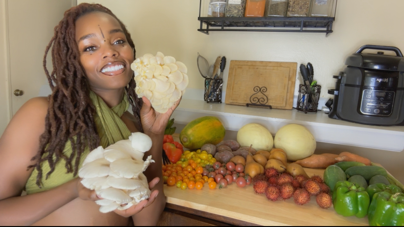 Woman with dreadlocks holding white mushrooms in a kitchen with vegetables on the counter.
