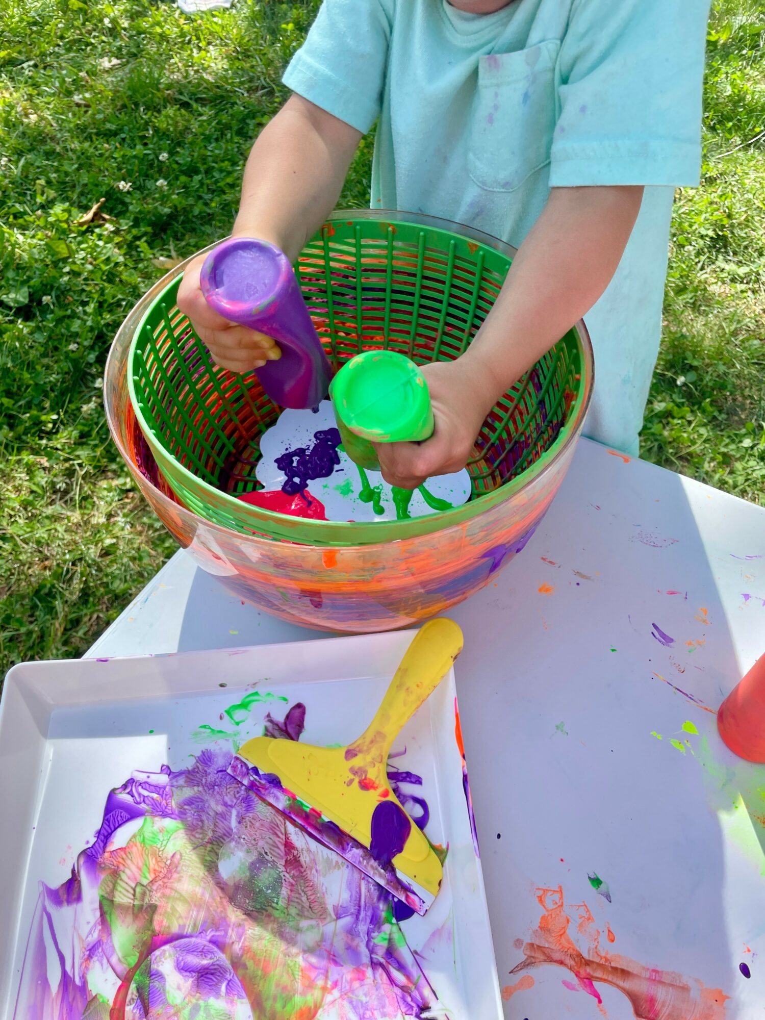 Child pouring purple and green paint into a basket with water outdoors, with paint on paper and tray nearby.