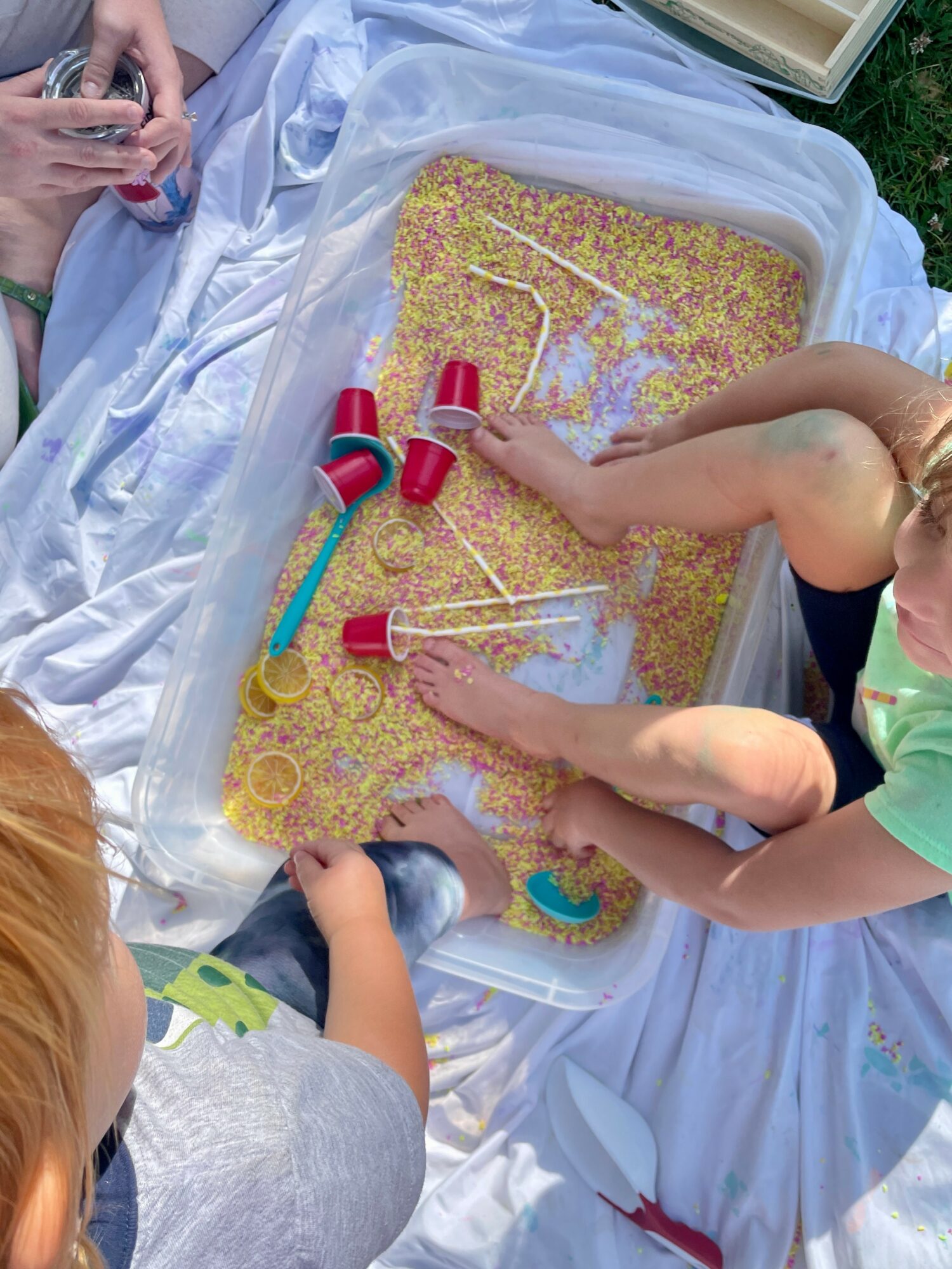 Children playing with a sensory water bead activity outdoors on a white cloth, with colorful tubes and sticks.